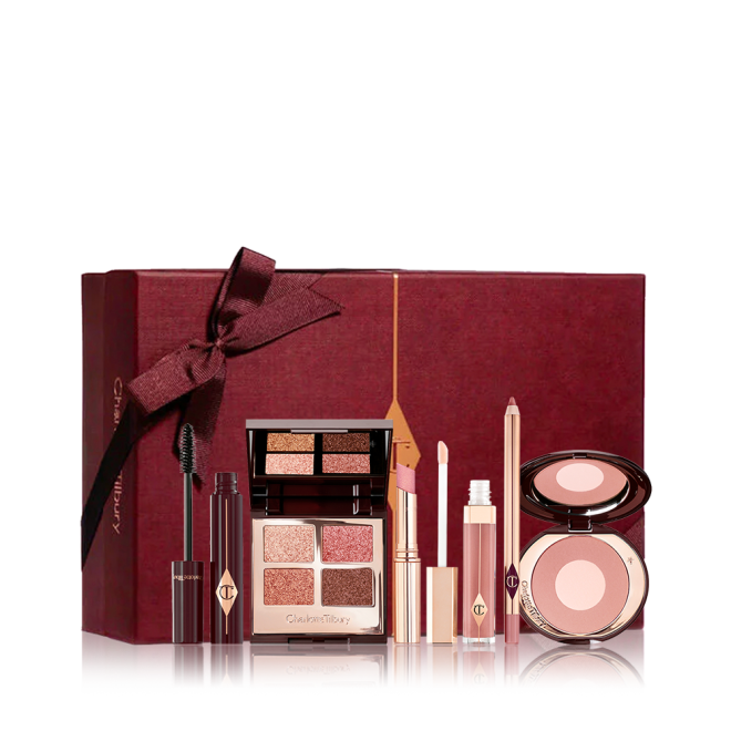 An open mascara tube with the applicator next to it with an open, mirrored-lid quad eyeshadow palette in shimmery neutral shades, a shimmery nude pink lipstick, a nude pink lip gloss and lip liner pencil, and a two-tone powder blush compact in a nude pink shade, with all products in front of a maroon-colour gift box. 