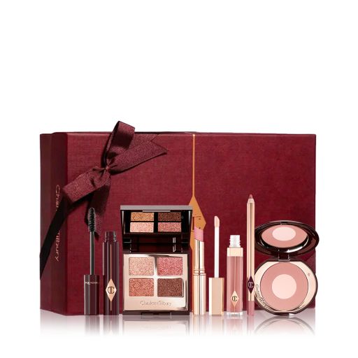 An open mascara tube with the applicator next to it with an open, mirrored-lid quad eyeshadow palette in shimmery neutral shades, a shimmery nude pink lipstick, a nude pink lip gloss and lip liner pencil, and a two-tone powder blush compact in a nude pink shade, with all products in front of a maroon-colour gift box. 