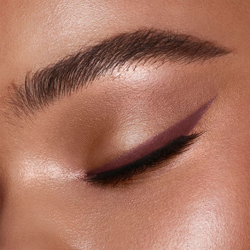 Close-up of a deep skin model with glowing skin and groomed eyebrows wearing a berry-brown eyeliner in a soft winged shape. 