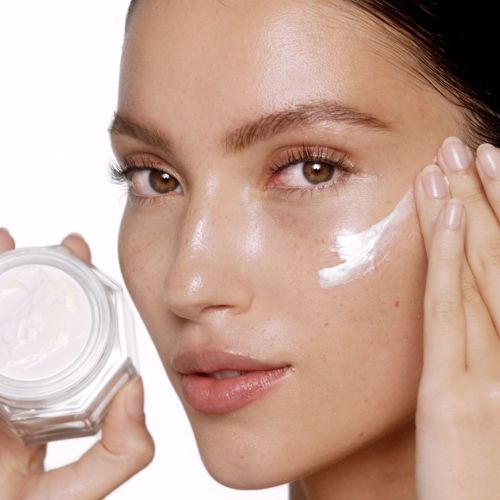 Fair-tone brunette model applying pearly-white face cream from a glass jar on her luminous, fresh-looking skin. 