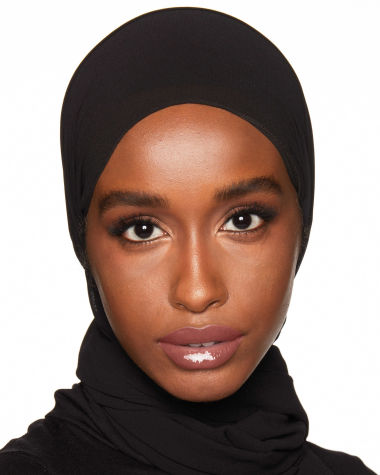 Close-up of a deep-tone model with a head-covering wearing a high-shine, sheer lip gloss in a nude-pink shade. 