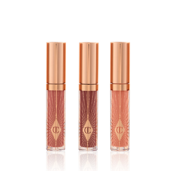 Three, travel-size lip glosses in shades of light pink, coral-peach, and brown-pink in glass tubes with gold-coloured lids. 