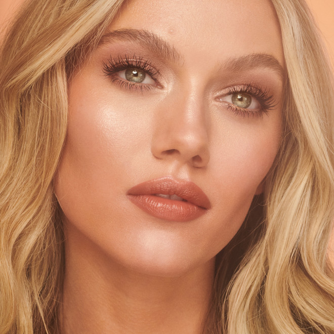Fair-tone model wearing glowy, flawless face base with shimmery eye makeup, and a rosy terracotta coral lipstick with a satin-finish.