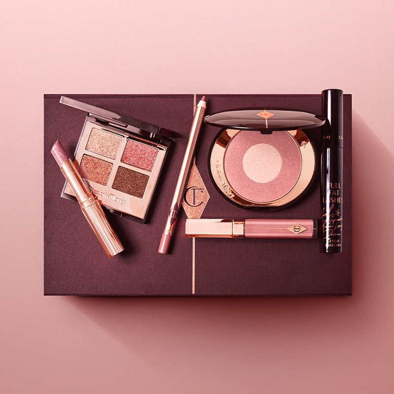 A mascara tube with an open, mirrored-lid quad eyeshadow palette in shimmery neutral shades, a shimmery nude pink lipstick, a nude pink lip gloss and lip liner pencil, and a two-tone powder blush compact in a nude pink shade, with all products placed on a maroon-colour gift box. 