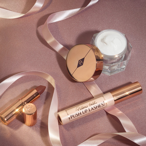 Pearly-white face cream in an open glass jar with a gold-coloured lid, black mascara in a pink tube with a gold-coloured lid, and golden-peach matte lipstick in a gold-coloured tube.