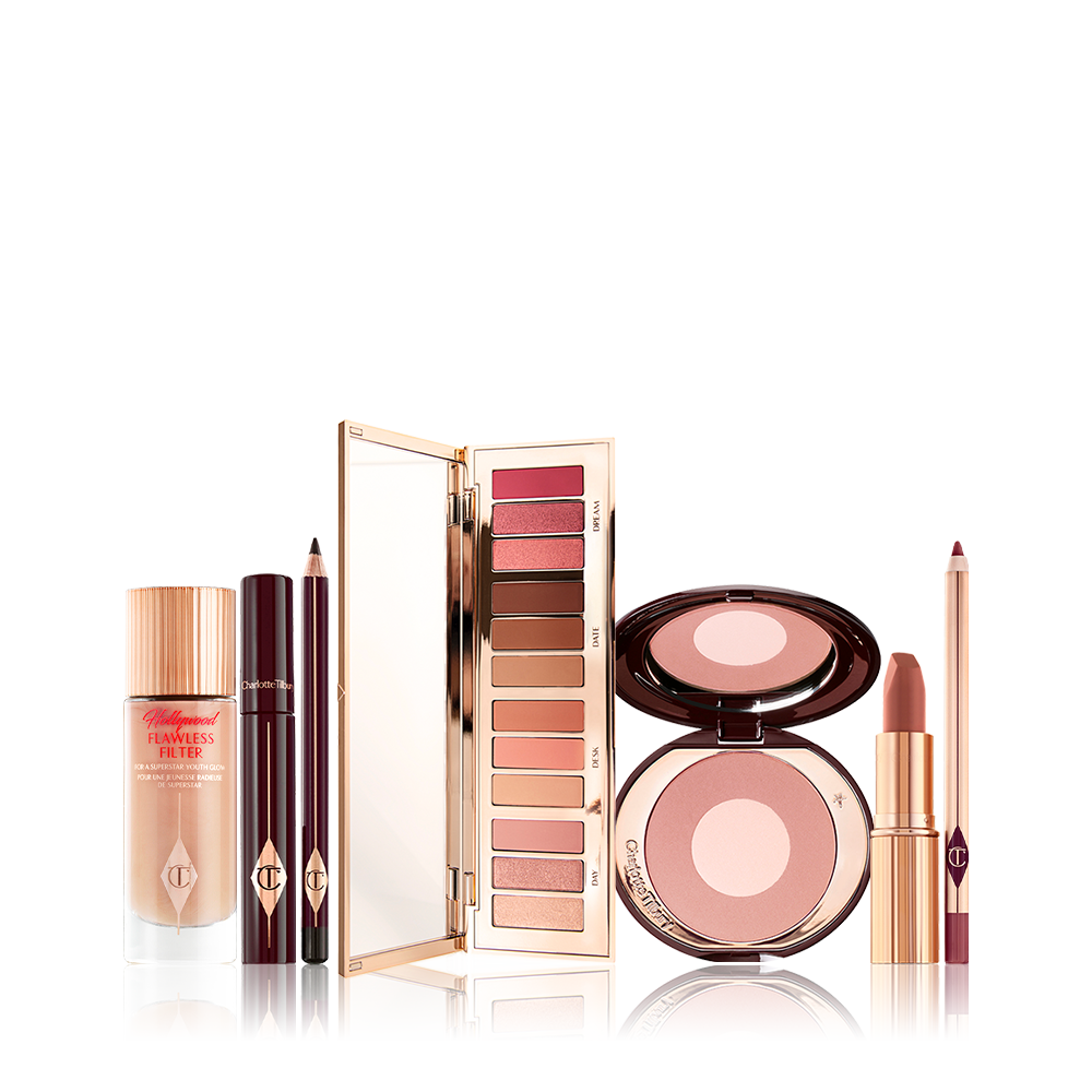 An open eyeshadow palette in pink, peach, gold, and brown shades, luminous primer in a glass bottle, black mascara and eyeliner, two-tone blush in light pink, and lip liner and lipstick in nude pink. 