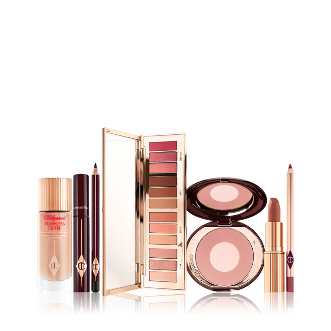 An open eyeshadow palette in pink, peach, gold, and brown shades, luminous primer in a glass bottle, black mascara and eyeliner, two-tone blush in light pink, and lip liner and lipstick in nude pink. 