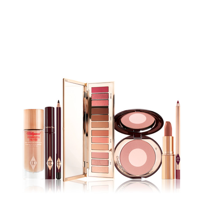 An open eyeshadow palette in pink, peach, gold, and brown shades, luminous primer in a glass bottle, black mascara and eyeliner, two-tone blush in light pink, and lip liner and lipstick in nude pink. 