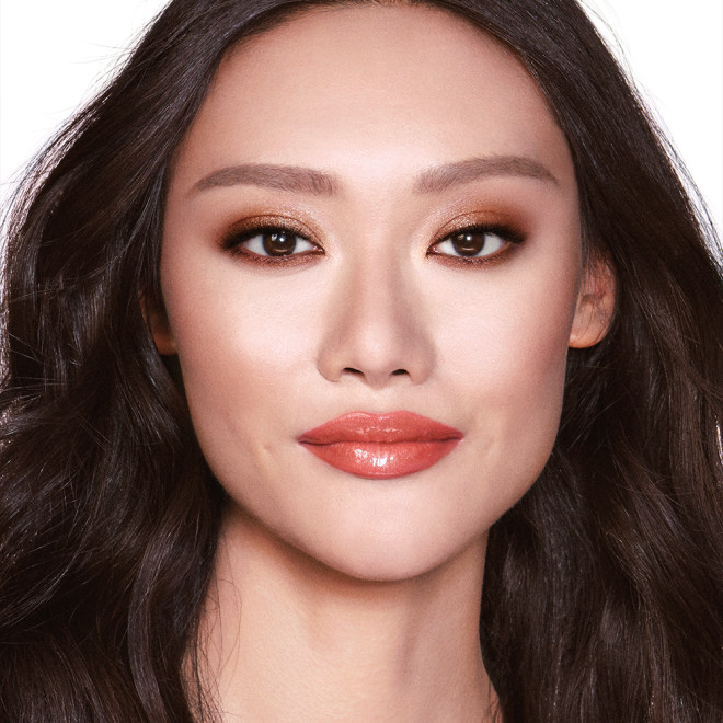 A fair-tone model with brown eyes wearing smokey brown eye makeup with warm bronze and pink blush, and glossy terracotta lips.