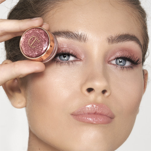 Fair-tone model with blue eyes wearing shimmery rose gold eye makeup and applying a pink glittering lip gloss with rose gold sparkle, while holding the rose gold eyeshadow pot in front of her.
