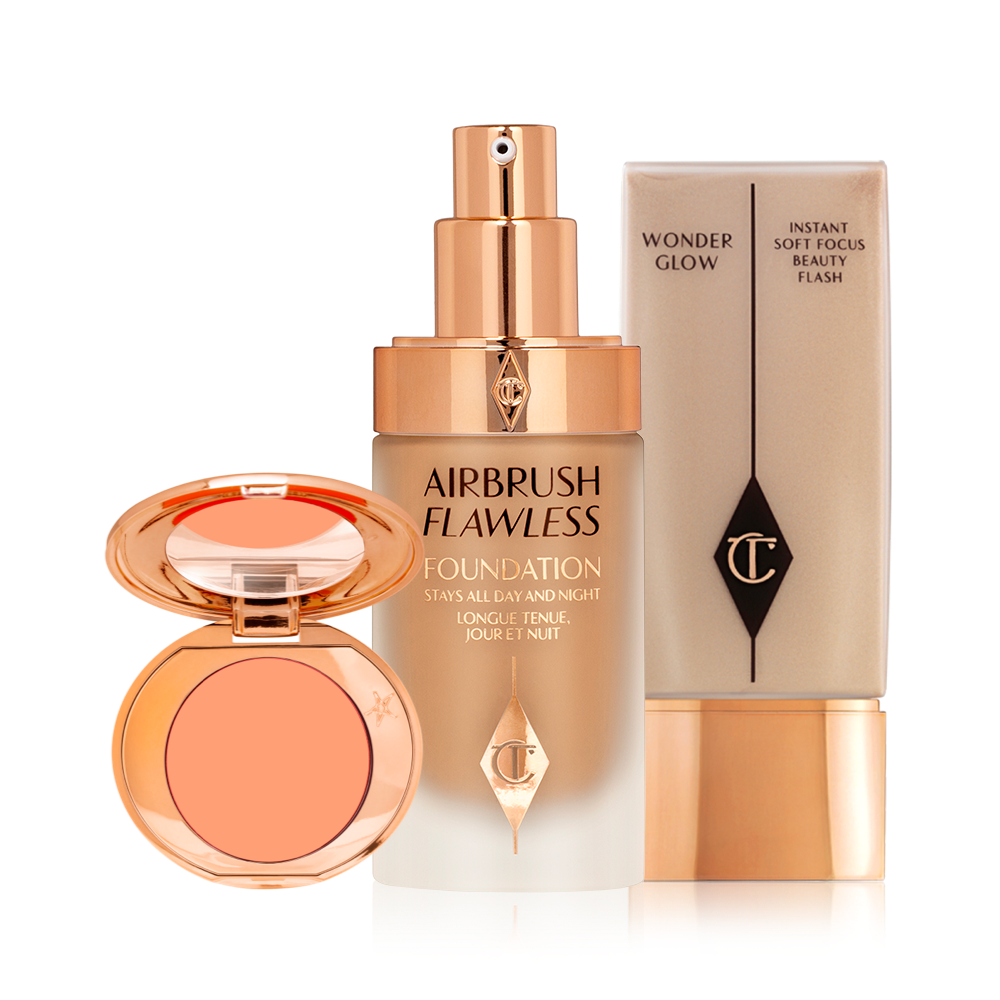 An blush compact with a mirrored lid with a peach shade with an open foundation in a glass bottle with a gold pump dispenser and a glowy primer in a clear bottle with gold-coloured lid. 
