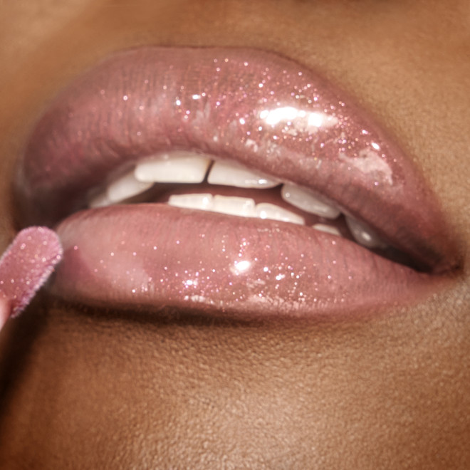 Lips close-up of a deep-tone model applying a nude pink lip gloss with a rose-golden sparkle.