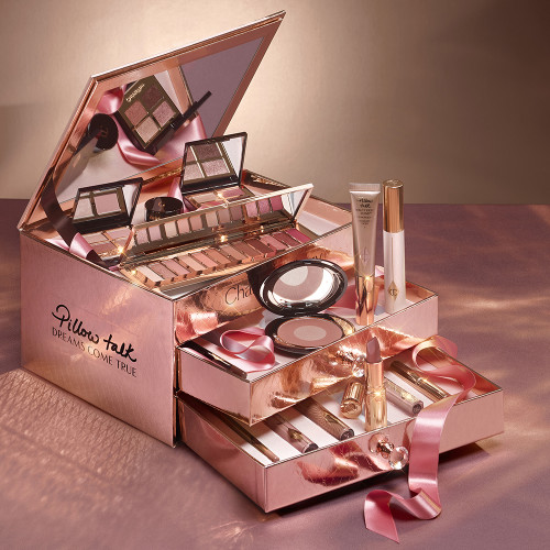A reflective, gold-coloured chest with two, opened drawers containing makeup items, which include Liquid highlighter wand in rose gold, black mascara, shimmery lip gloss in nude pink, glitter-free but high-shine lip gloss in nude pink, rose gold eyeshadow pigment in a petite pot, two closed lipsticks, travel-size lip gloss in nude pink, eyeliner pencil in dark brown, lip liner pencil in nude pink, quad eyeshadow palettes, 12-pan eyeshadow palette, and two-tone blush compacts.