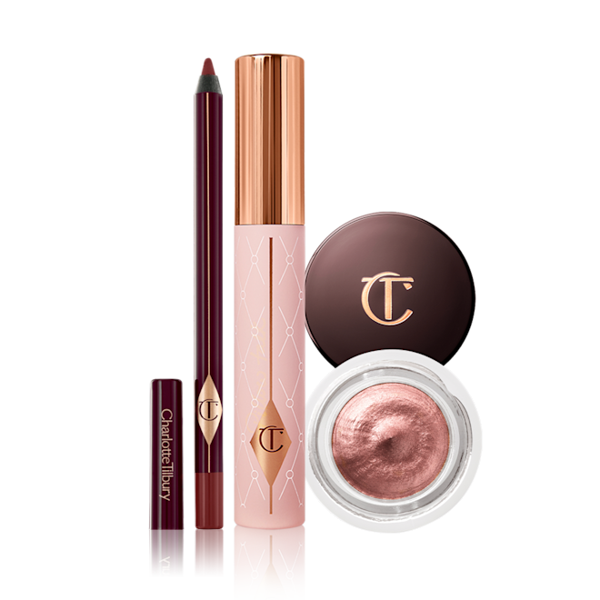 Charlotte's Mesmerise Define Eye Kit open An open eyeliner pencil in a berry-brown shade, mascara in a nude pink tube with a gold-coloured lid, and cream eyeshadow in a rose gold shade in a glass pot with a dark-brown-coloured lid.