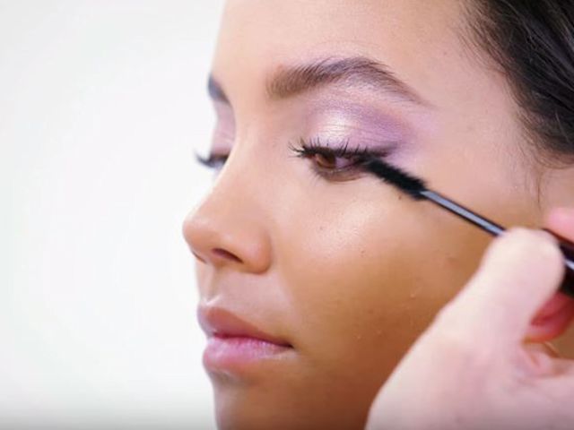 A light-tone model wearing frosted lilac eye makeup and getting a black, volumising mascara applied. 