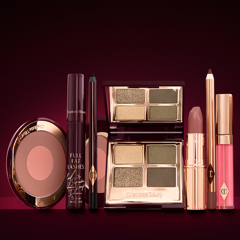 An open two-tone blush in cool-toned brown and warm pink with a mascara, eyeliner pen, quad eyeshadow palette with shimmery and matte green and golden shades, an open lipstick in nude red-brown, lip liner pencil in taupe-brown, and a lip gloss in bright pink. 