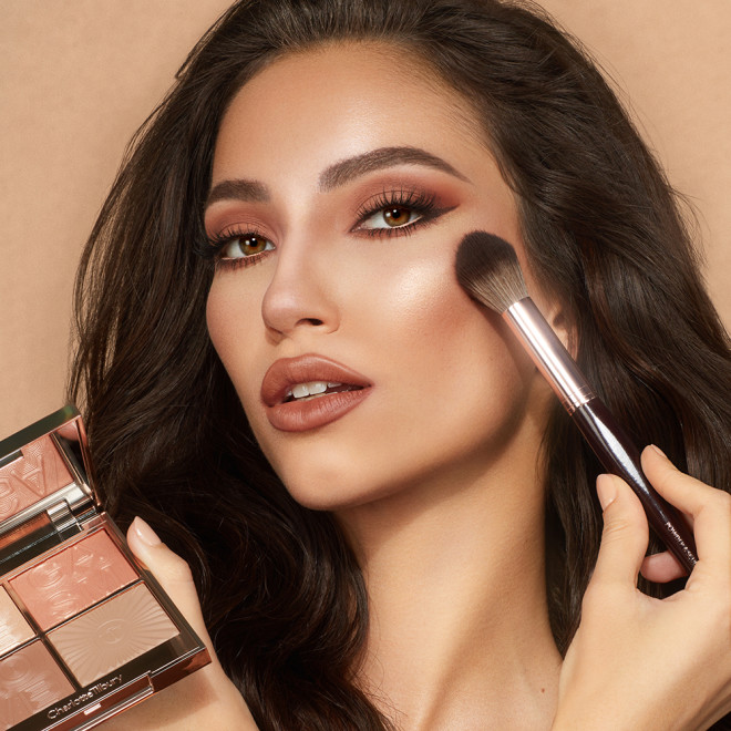 Medium-tone brunette model with brown eyes and glowy, bronzed skin, applying peach highlighter blush from a quad face palette with contouring, eyeshadow, and highlighter in nude shades.