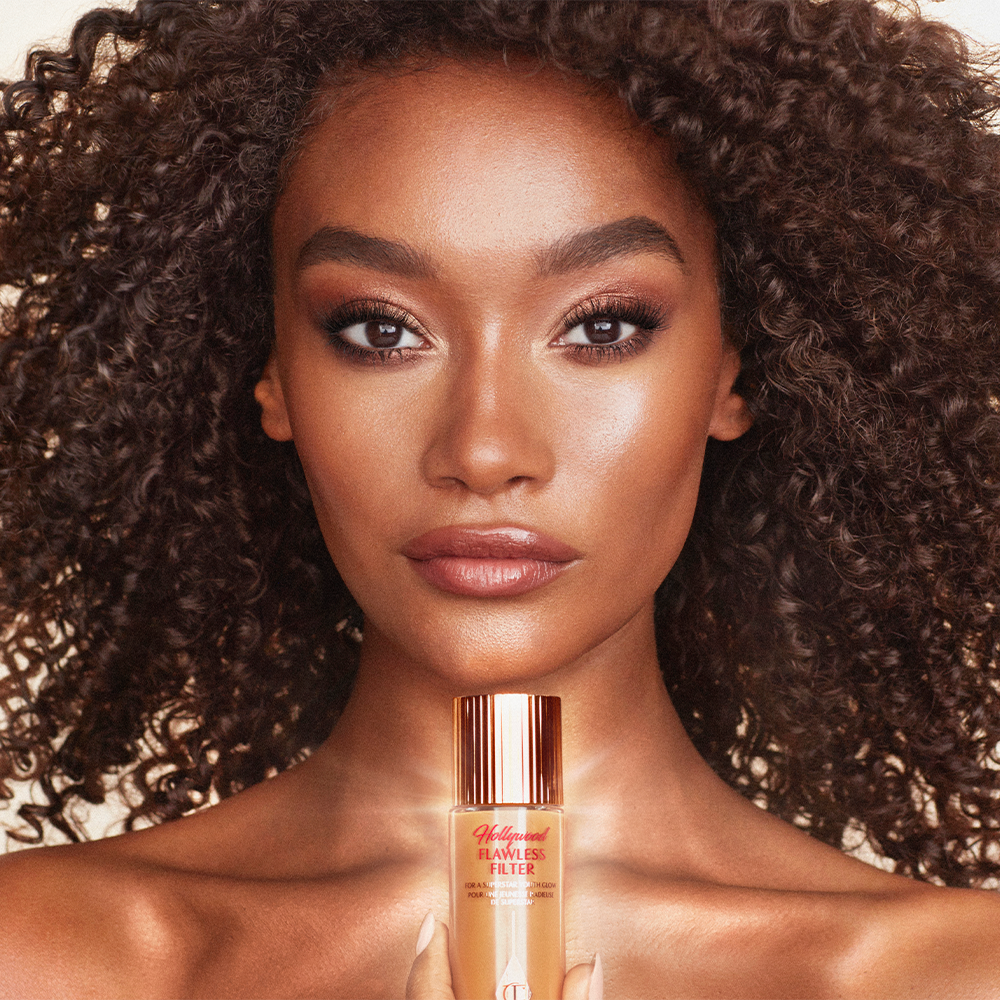 Deep-tone model with glowy, flawless skin holding a luminous primer in a medium shade in a glass bottle with a gold-coloured lid. 