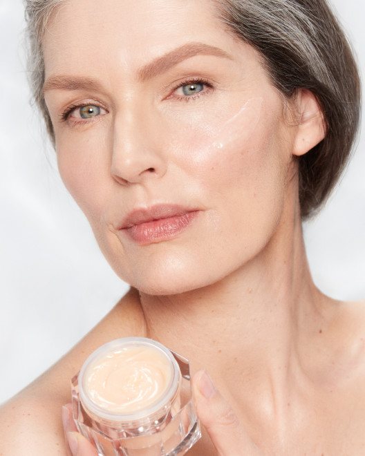 Fair-tone model with glowy, fresh, and mature skin, holding an open glass jar of a thick, peach-coloured night cream.