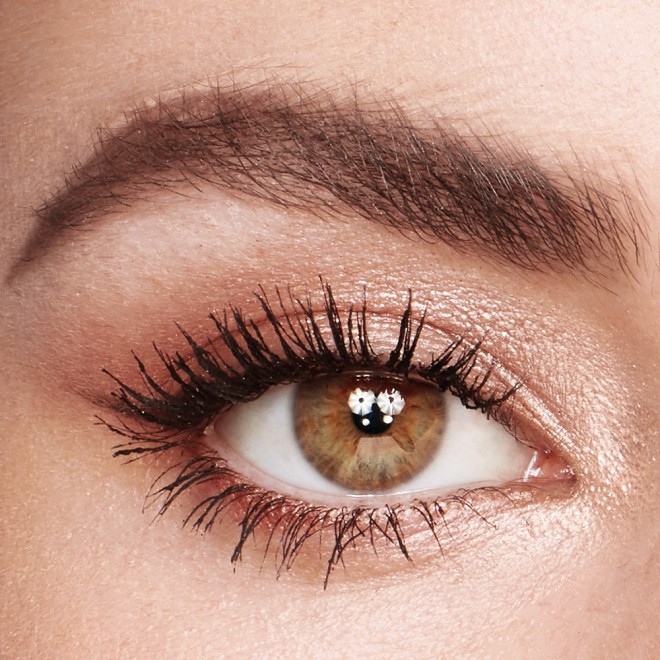 Single-eye close-up of a fair-tone model with hazel eyes wearing shimmery champagne, light brown, and rose gold eye makeup with jet-black lengthening mascara.