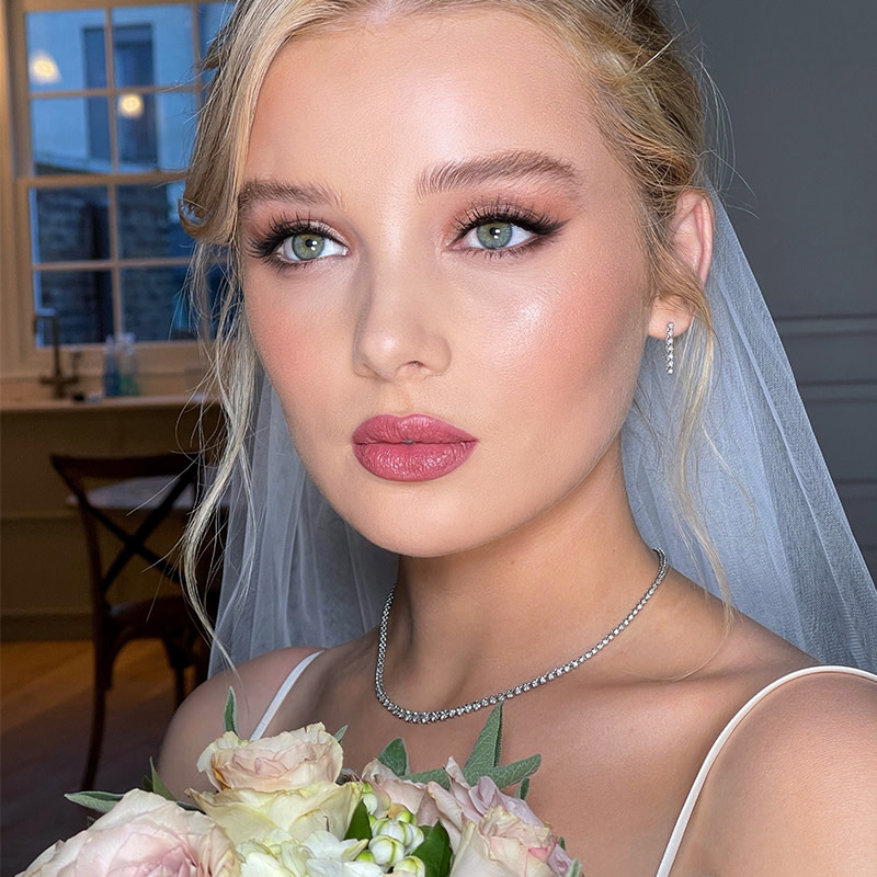 Fair-tone blonde bride with blue eyes wearing a glowy, fresh makeup base with coral blush, dark berry-pink lips, and shimmery fawn-coloured eye makeup with black eyeliner.