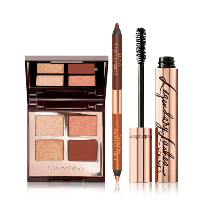 An open, mirrored-lid, quad eyeshadow palette with eyeshadows in shades of copper and brown with an open, double-ended eyeliner in copper and champagne, and a black mascara with its applicator next to it. 