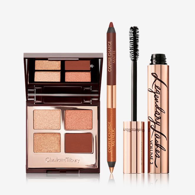 Copper Charge Magic Trick Eye Kit open An open, mirrored-lid, quad eyeshadow palette with eyeshadows in shades of copper and brown with an open, double-ended eyeliner in copper and champagne, and a black mascara with its applicator next to it.