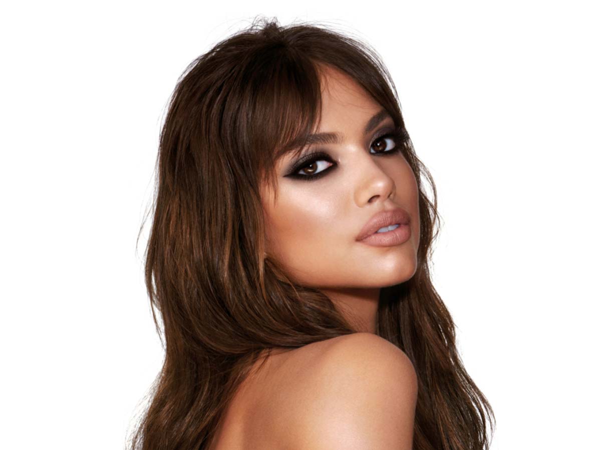 A medium-tone brunette model with brown eyes wearing smokey silvery eyeshadow with bronzed cheeks and glossy nude-peach lips. 