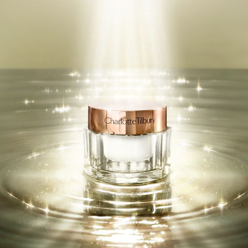 A pearly-white face cream in a glass jar with a rose gold coloured lid, on top of glimmering water with light shining on it. 