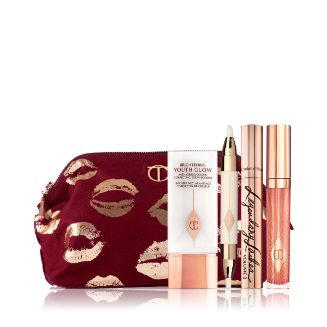 Primer in a white bottle with a rose gold-coloured lid with a double-ended concealer pen, mascara in gold-coloured tube, and a nude pink lip gloss with a gold-coloured lid along with a red makeup bag with a kiss prints all over it.