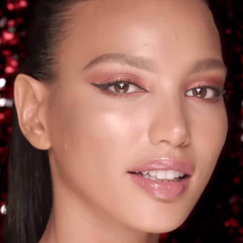 A medium-tone model with brown eyes wearing bright rose-gold eye makeup, black eyeliner, and nude pink lip gloss. 