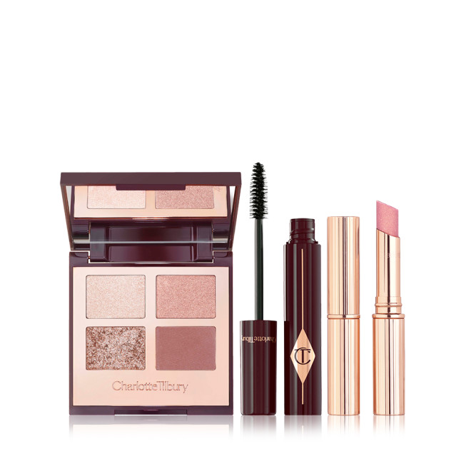 An open, mirrored-lid quad eyeshadow palette in nude shades, a mascara in a dark crimson and gold colour with its applicator next to it, and a sparkly, nude pink lipstick. 