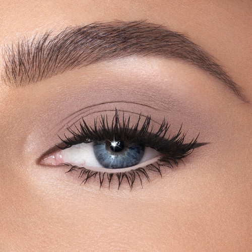 Single-eye close-up of a fair-tone model with blue eyes wearing dark beige cream eyeshadow smudged and spread on her eyelid with black eyeliner for a smokey beige eye look. 