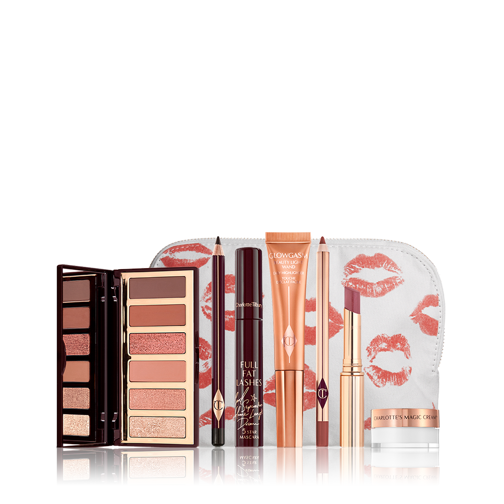An open, mirrored-lid eyeshadow palette in nude brown and peach shades, an open eyeliner pencil in black, mascara, highlighter wand in peach packaging, lip liner pencil in redwood, lipstick lip balm in sheer red, and mini face cream in a glass pot, with all products displayed in front of a white-coloure makeup pouch. 