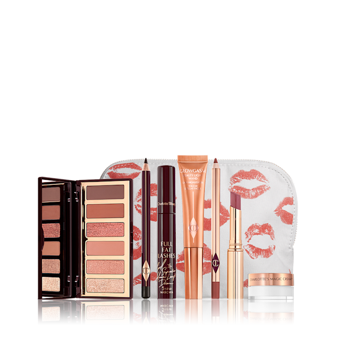 The Charlotte Darling kit with Mini Magic Cream displayed An open, mirrored-lid eyeshadow palette in nude brown and peach shades, an open eyeliner pencil in black, mascara, highlighter wand in peach packaging, lip liner pencil in redwood, lipstick lip balm in sheer red, and mini face cream in a glass pot, with all products displayed in front of a white-coloure makeup pouch.