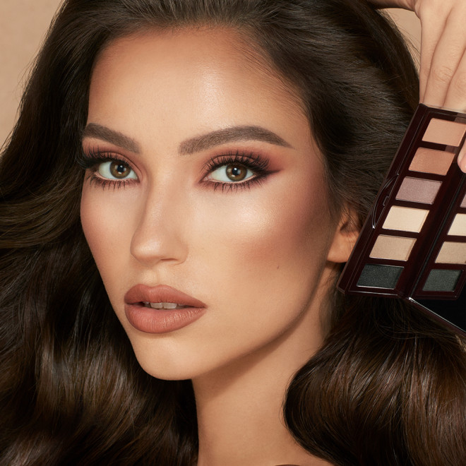 A medium-tone model wearing glowy nude brown makeup holding an open, mirrored-lid six-pan eyeshadow palette with matte eyeshadows in brown, peach, and beige shades.