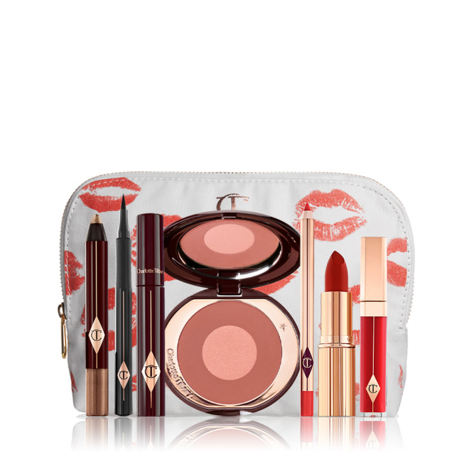 A white-coloured makeup pouch with an open two-tone blush in cool-toned terracotta and warm pink with a mascara, eyeliner pen, an open lipstick in bold red, lip liner pencil in blood-red, and a lip gloss in bright red. 