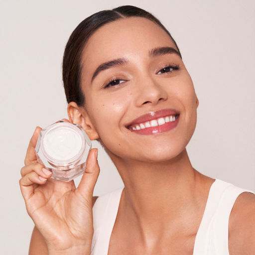 A medium-tone brunette model holding an open jar of thick, pearly-white cream. 