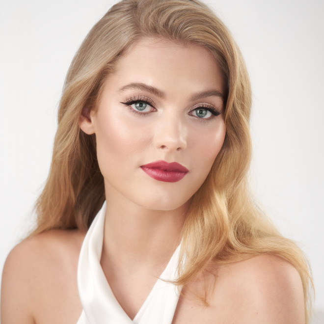 A fair-tone blonde model with blue eyes wearing soft, fawn eye makeup with glowy nude blush, and berry-rose matte lipstick.