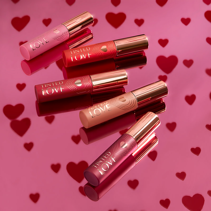 collection of lip and cheek tints placed on a heart-covered wallpaper in shades of pink, dark red, vibrant red, nude brown, and peachy-brown. 