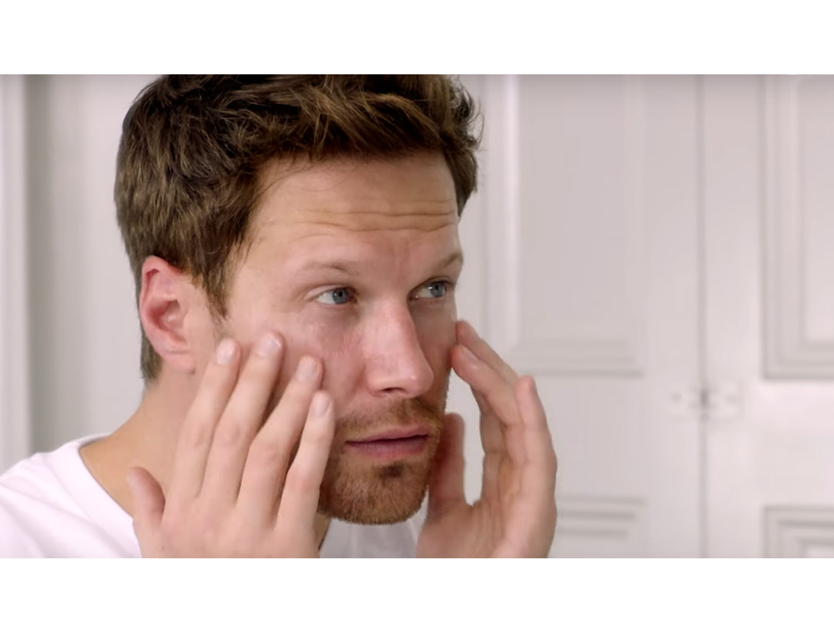 A light-tone, brunette male model patting a face cream into the skin. 