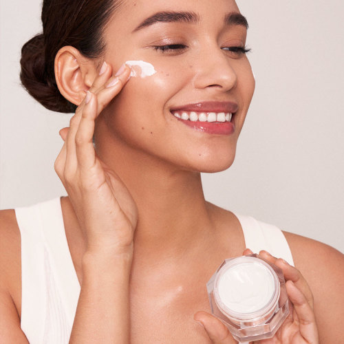 A medium-tone brunette model applying a thick, pearly-white cream from an open jar.