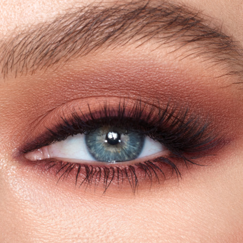 Single-eye close-up of a fair-tone model with blue eyes wearing smokey pink and brown eye makeup with champagne, highlight applied on her inner eye corner with lengthening black mascara.