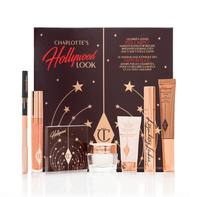  A double-ended eyeliner pencil in black and nude beige colours, high-shine lip gloss in a peachy-rose colour, quad eyeshadow palette with a brown-coloured lid, glowy, pearly-white face cream, liquid highlighter wand in rose gold, black mascara in a gold-coloured tube, and luminous primer, along with their packaging box.
