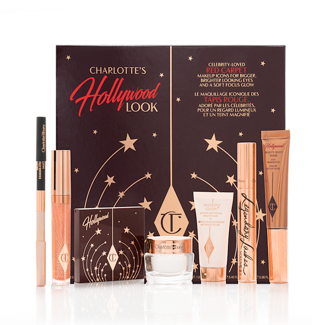 Charlotte's Hollywood Makeup Look with box A double-ended eyeliner pencil in black and nude beige colours, high-shine lip gloss in a peachy-rose colour, quad eyeshadow palette with a brown-coloured lid, glowy, pearly-white face cream, liquid highlighter wand in rose gold, black mascara in a gold-coloured tube, and luminous primer, along with their packaging box.