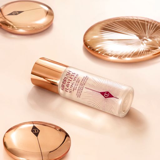 A setting spray in a clear bottle with a gold-coloured lid placed on a champagne-coloured floor with closed face powder and bronzer compacts all around.