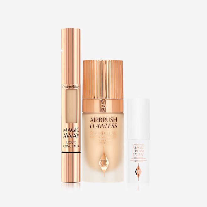 Charlotte's Magic Airbrush Complexion Kit displayed Creamy liquid concealer in a gold-coloured tube with a soft sponge applicator end, foundation in a frosted glass bottle with a gold-coloured lid, and pearly-white face cream in a glass jar with a gold coloured lid.