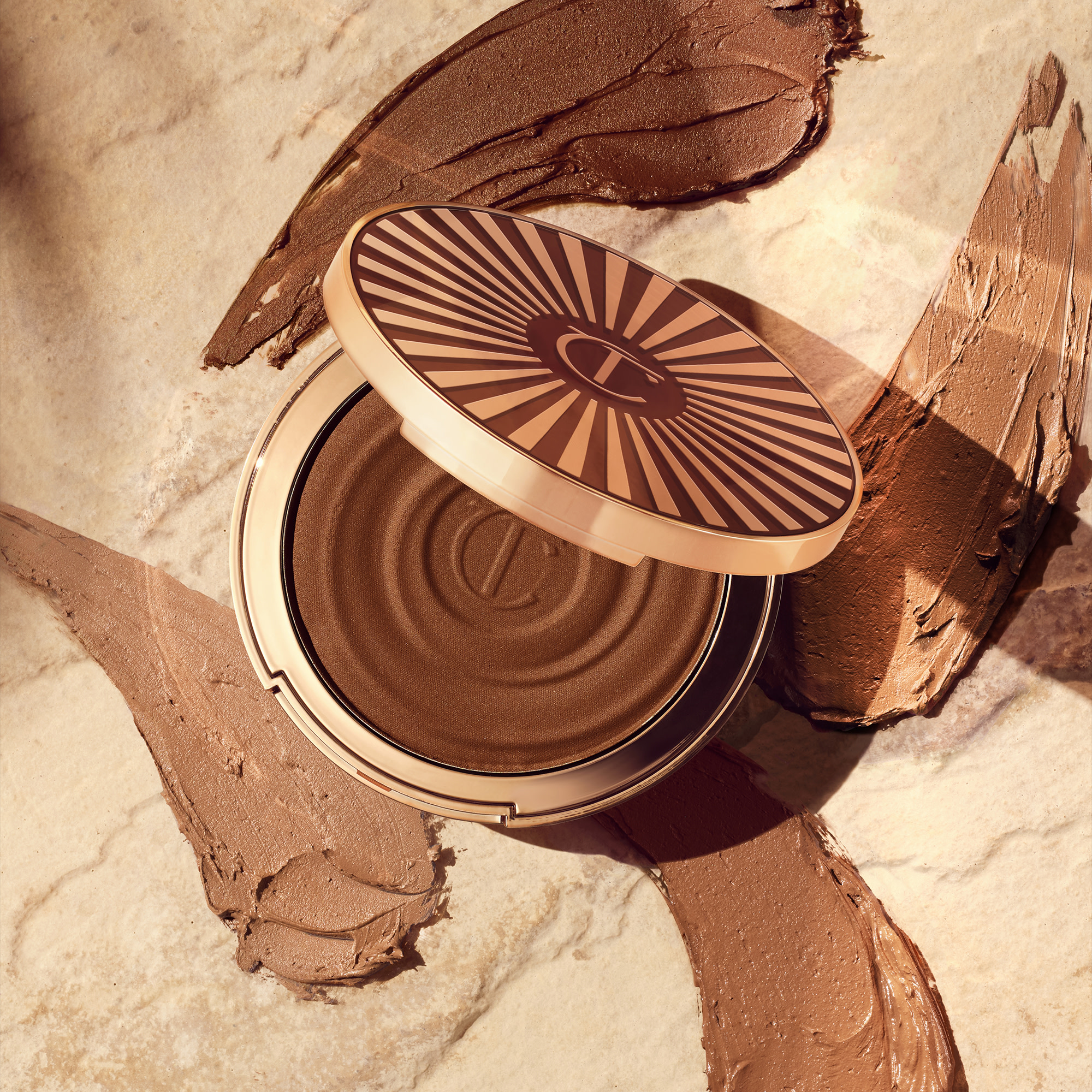 Open, cream bronzer compact in a light-sandy-brown shade with gold-coloured packaging with swatches of four different bronzers in various shades of brown behind it.