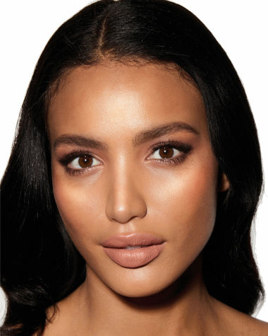 Deep-tone model with brown eyes wearing a nude peach matte lipstick with a glowy yet subtle, pearlescent highlighter in a rose-gold shade.