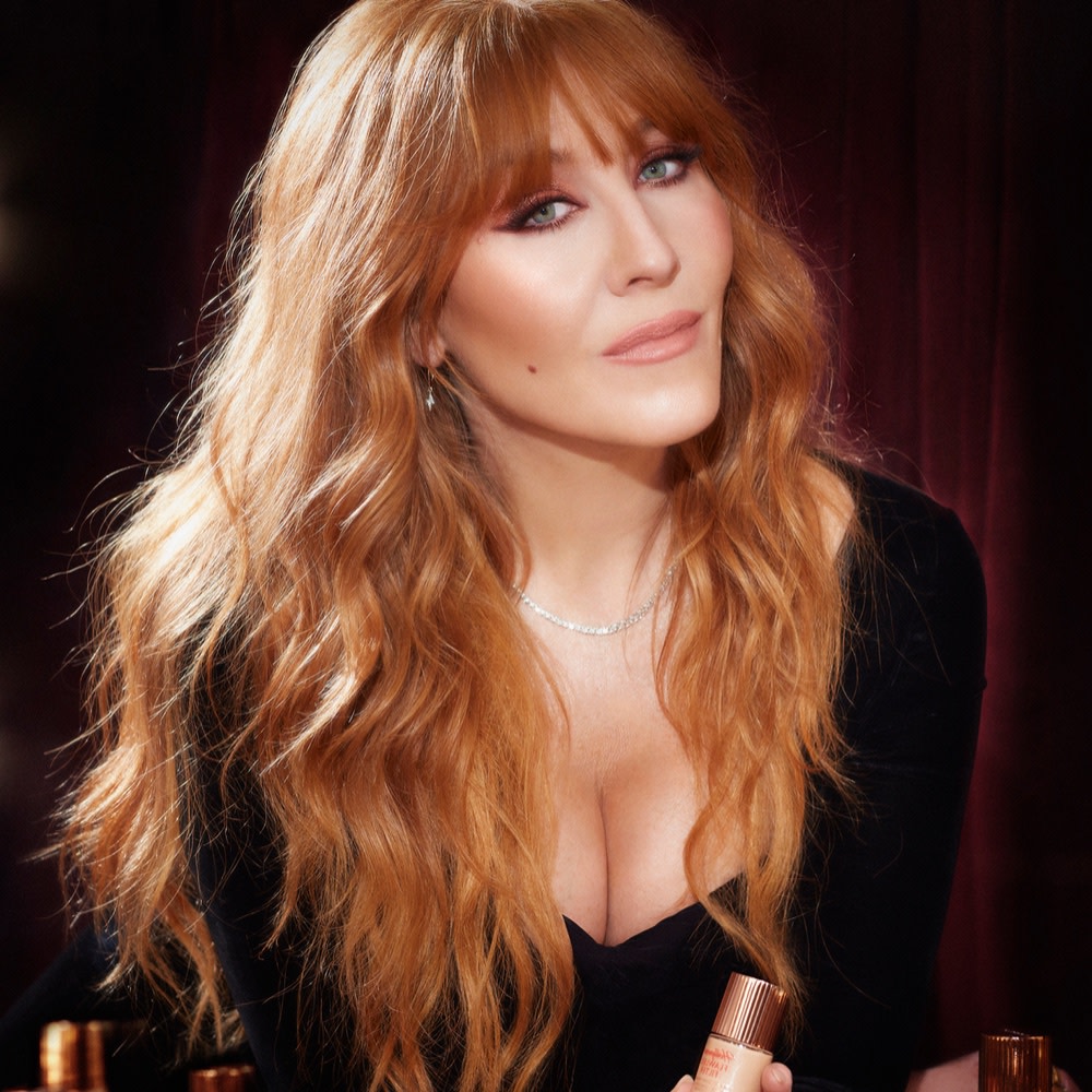 Portrait of Charlotte Tilbury with bronzed skin, wearing glowy, nude pink makeup, dangly earrings, and a black dress. 